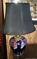 Full view of the table lamp showing the black fabric shade, brass fixture, and the cobalt blue pottery base decorated with large colorful flowers.
