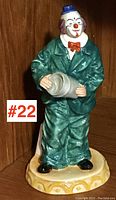 Front view of Royal Doulton clown figurine with green suit, red bow tie, blue hat, gray top hat in hands, standing on yellow decorative base.