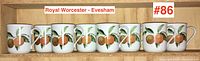 Eight white porcelain mugs with orange fruit design and gold rims on wooden shelf, front view