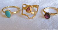Photo showing 3 gold-tone gemstone rings side by side on white surface featuring turquoise, garnet, and amethyst stones.
