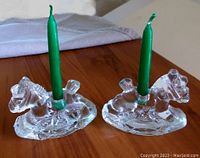 Two solid glass rocking horse candle holders each fitted with a green candle, placed on wooden surface