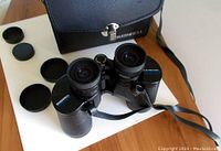 Bushnell binoculars with lens caps off, showing lens details and strap, placed on table with carrying case behind.
