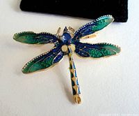 Close-up photo of dragonfly brooch showing detailed blue and green enamel wings, gold-tone metal frame with beaded edges and large blue jewel as head.