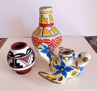 Three Southwestern decorative pottery pieces shown on flat white surface: round patterned vase, multi-tiered vessel with geometric designs, bird-shaped flower painted vessel.