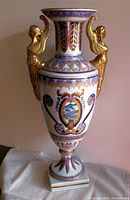 Front view of the vintage porcelain urn showing the hand-painted motifs, gilding, scenic medallion, and winged Victory handles.