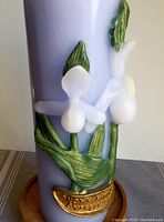 Close-up side view of pale blue pillar candle with white wax lady slipper orchid flowers, green leaves, and gold wax banner with embossed text on wooden base.
