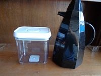 Photo showing black geometric coffee dispenser with handle, and clear square plastic food storage container with white lid