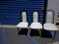 Three metal dining chairs with black frames and white vinyl padding, placed outdoors on pavement with blue storage unit doors in background.