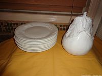 Stack of 8 white small plates with embossed fruit and vine design around edges, next to a large white ceramic pear figurine.