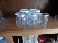 Two round glass storage jars with clear plastic lids alongside one round plastic container.