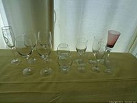 Photo showing all 12 glassware pieces arranged in a row on a yellow cloth, varied shapes including wine glasses, snifter, and pink tinted glass.