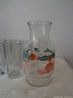 Side view of a clear glass jar decorated with colorful citrus fruit graphics and seven matching clear drinking glasses with ribbed texture.
