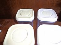 Top view of four square or rectangular plastic storage container lids in white and light purple showing oval seals.