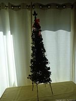 Full view of the slender artificial Christmas tree showing its height and leafy texture against light-colored curtains.