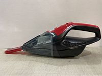 Side view of the red and black Dirt Devil cordless handheld vacuum cleaner with transparent dust container and red quick-flip crevice tool attached.
