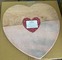 Heart-shaped serving board showing the natural pinkish marble and acacia wood materials, placed on a cardboard box with a heart-shaped label attached at center.