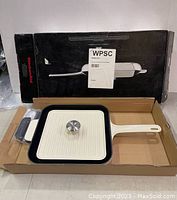 KitchenAid cast iron grill pan with lid, placed inside original cardboard packaging with handle covers and instruction manual included