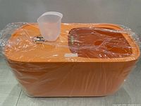 Full view of the orange plastic rice container wrapped in clear plastic, showing the rectangular shape and included measuring cup on top.