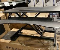 Side view of the black standing desk converter showing the dual tiers and hydraulic lift mechanism, placed on a cardboard box with product illustration.