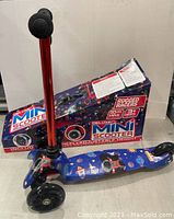 Front angle view showing the space-themed deck, front wheels, and partially visible adjustable handlebars with original box in the background.