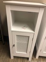 Front view of the white bathroom cabinet showing the woven panel door and top shelf space with small hardware piece inside.