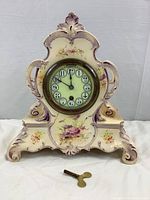 Front view of the ceramic mantel clock with floral hand painted design, showing overall shape and winding key placed in front.