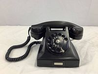 Black Bakelite rotary dial telephone shown from front with handset on cradle and rotary dial visible.