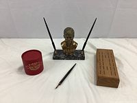 Wide shot of all three items: brass JFK bust pen stand with two black pens, red ink bottle box, and wooden cribbage board with pegs.