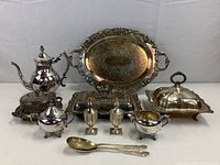 Photo showing full set of silver plated items arranged on and around an ornate silver tray. Items include coffee pot with burner stand, covered dishes, serving spoons, salt and pepper shakers, creamer, sugar bowl, and lids.