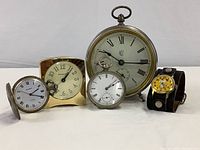 All five timepieces displayed together on white surface showing various sizes and styles including Tiffany & Co. desk clock, 2 pocket watches, Snoopy wristwatch, and Waterbury alarm clock