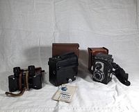 Three vintage optical devices arranged side-by-side on a white cloth background. From left to right: Ottowalter binoculars with case, black Spartus press flash camera with case, Ciro-flex twin-lens reflex camera in leather case with shoulder strap, and Spartus flash bulb packet placed in front.