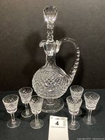 Front view of decanter with stopper and six matching cordial glasses against black background