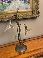 Full view of the art glass table lamp with two curved glass floral shades on twisted metal arms, placed on a wooden surface before a framed painting.