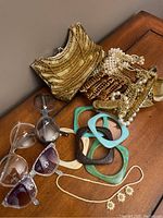Gold beaded handbag, several chunky vintage bracelets, faux pearl necklaces, and four pairs of sunglasses arranged on a wooden surface.