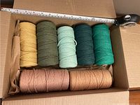 Top-down image of 7 spools of macrame cord in a cardboard box with measuring tape to show size, colors vary from yellow, green to peach and brown tones.
