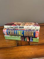 Stack of three cookbooks on a wooden surface: The Lodge Cast Iron Cookbook, The New Grilling Book, and The Barbecue Collection.