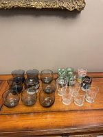 Full view of all 18 mid century vintage glasses arranged on wood surface, showing different styles and colors including smoked amber, green floral, clear floral, and decorative patterned glasses