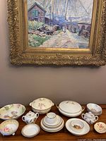 Overview of dish set displayed under painting showing covered bowls, plates, teacups, and sugar bowl with gold edges and floral patterns