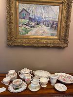 Full view of the gold and floral dish set including cups, plates, sugar bowl, creamer, and serving platters arranged on a wooden table under a framed painting.
