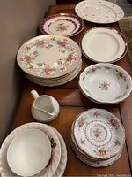 Display of assorted decorative plates stacked on a wooden table, showing various floral and gold patterned plates, a white creamer, and several bowls with no visible damage.
