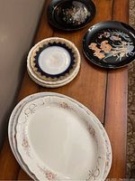 Photo showing black round plates with floral designs, blue and gold rimmed plates stacked, and floral gold-accented oval dish stacked.