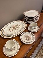 Photo showing stacks of round steak plates with 3 oval platters in front, plus 2 side plates, 2 cups and 2 saucers arranged on a wooden table.