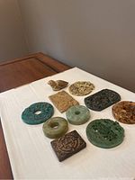 Overhead view of 10 carved jade and stone pendants on a white surface alongside a carved bird figurine pendant, showing a variety of shapes and colors from green, blue, beige to brown.