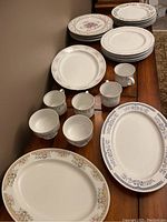 Assorted white ceramic mugs, plates, and platters on wooden table featuring floral and geometric designs, no visible chips or cracks.