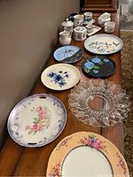Assorted decorative plates with floral and botanical patterns along with white mugs and lidded dishes with blue designs, displayed on wooden surface