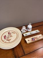Four round decorative plates with a brown botanical design, two rectangular dishes, a small white bell with floral decoration, and a 101 Dalmatians figurine on wooden surface.