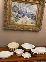 Photo showing seven pieces of Pyrex and ceramic bakeware under a gold framed painting on a wooden table.