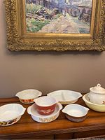 All bakeware pieces arranged on wooden surface under a painting, showing various colorful fruit and floral designs typical of vintage Pyrex sets.