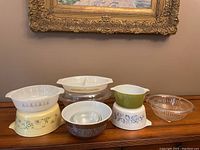 Photo showing entire collection of Pyrex and assorted glass bakeware arranged on wooden surface with framed painting in background.