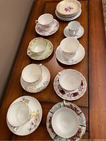 Overview of 9 teacups and 9 saucers arranged on wooden table, showing floral patterns in pastel and bright colors, some cups without saucers.